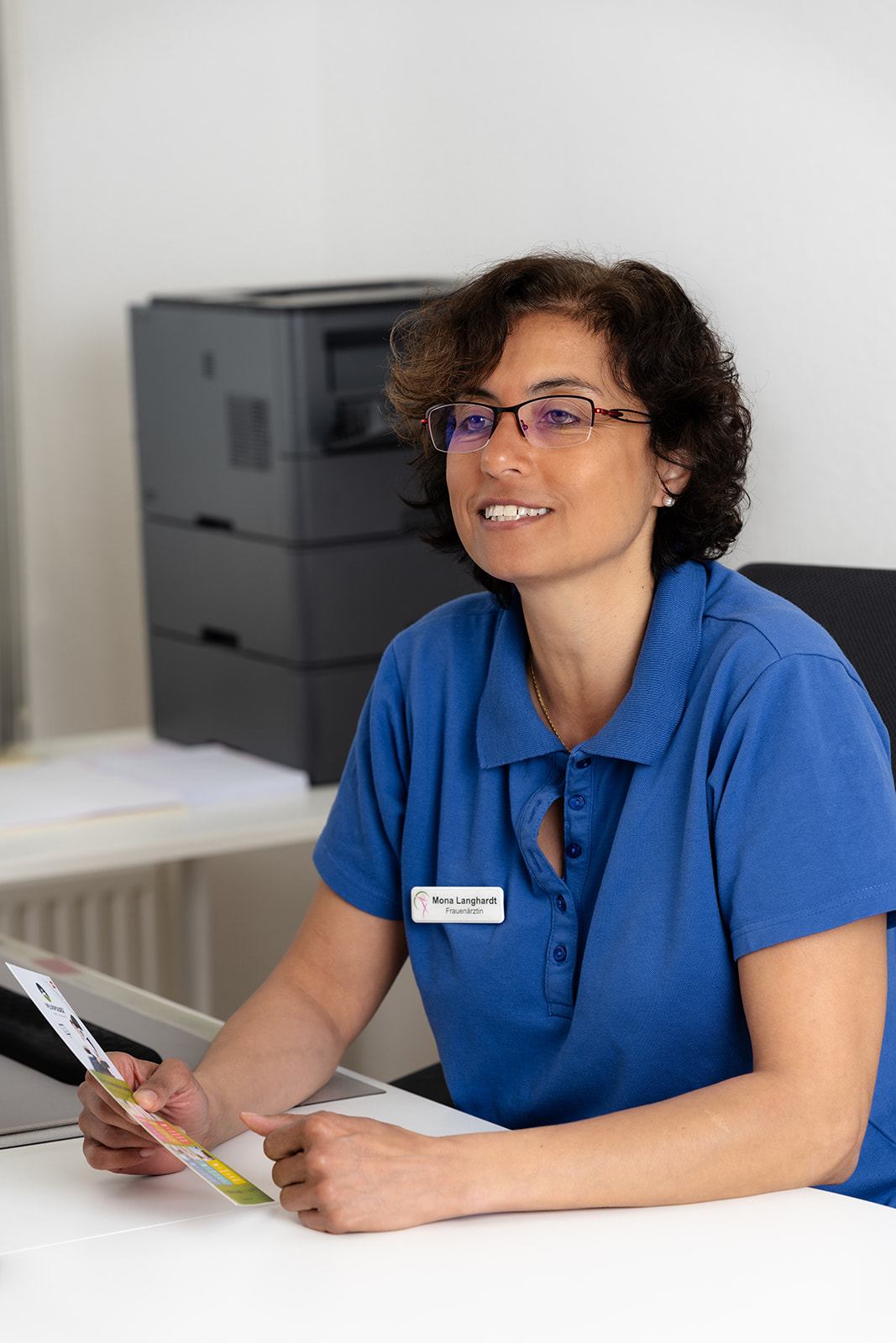 Frau Mona Langhardt sitzt mit einer Broschüre in der Hand am Schreibtisch und erklärt freundlich lächenld.